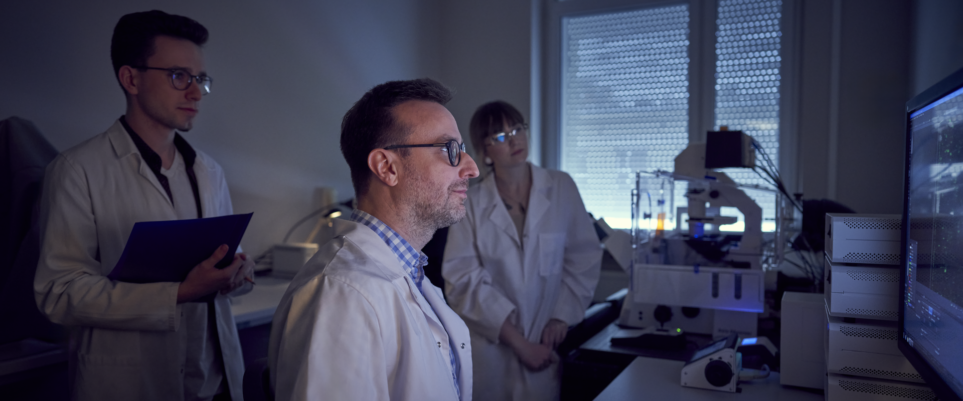 profesor siedzi przed komputerem w laboratorium, za nim stoi dwójka doktorantów, która przygląda się informacjom wyświetlanym na ekranie