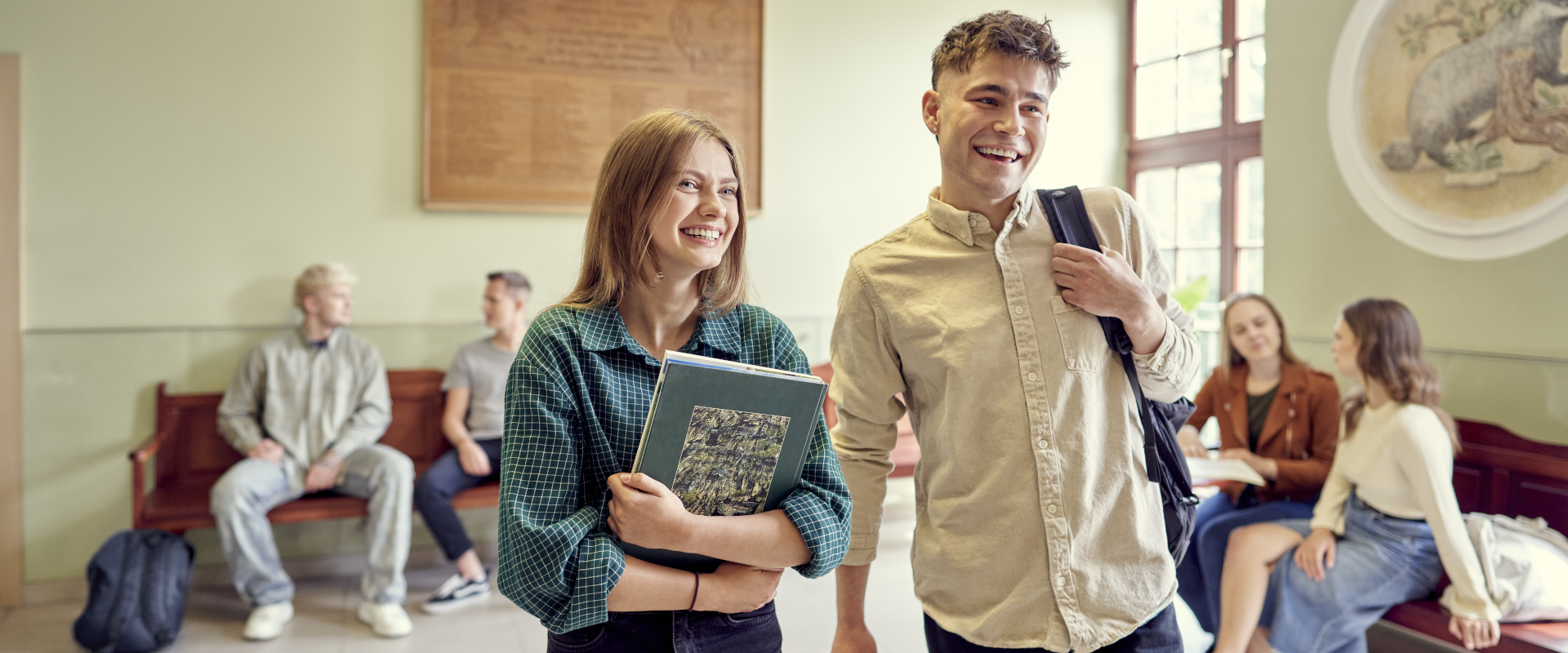 studentka i student idą uśmiechnięci z książkami i plecakami, na drugim planie inni studenci rozmawiają