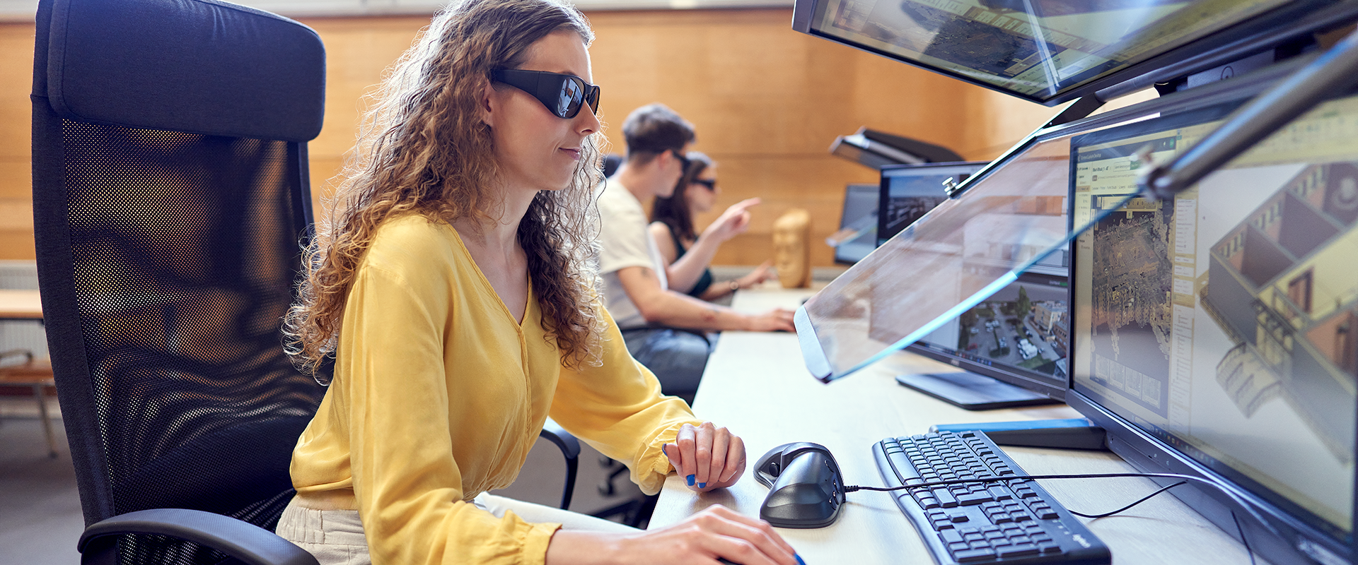 dziewczyna siedzi przy komputerze do pracy w 3D, ma na sobie specjalne okulary, w tle pracują dwie inne osoby