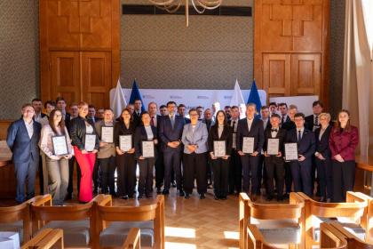 Grupa ludzi stoi w sali, trzymając certyfikaty w rękach, uśmiechając się do kamery.