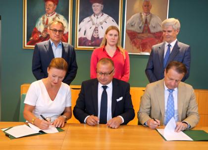 Trzy osoby podczas podpisywania dokumentów, w tle stoją trzy osoby